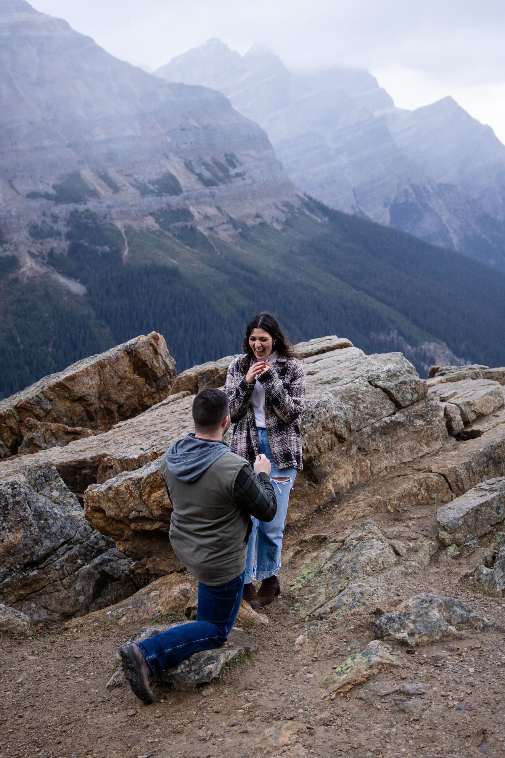 Surprise proposal in Banff with a man kneeling on a mountain overlook as his partner reacts with joy, surrounded by Banff National Park scenery.